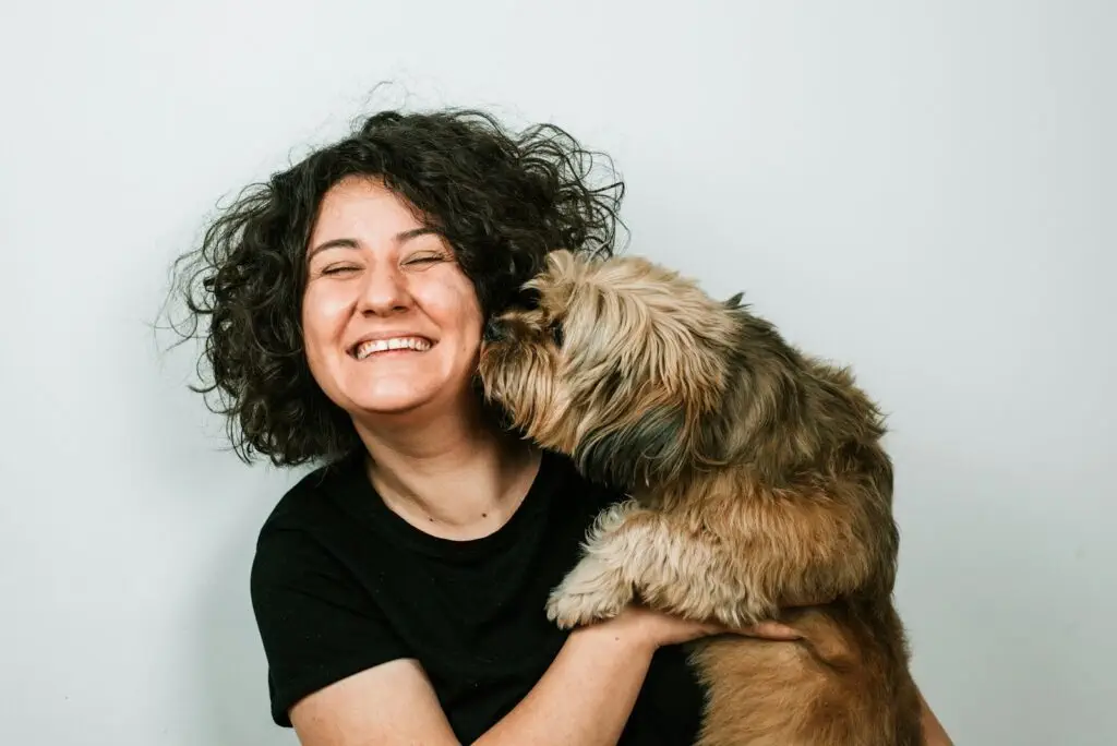 A dog kissing a woman with black curly hair who is wearing a black shirt.